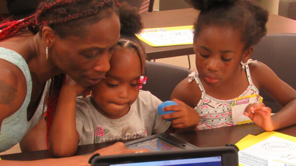 An adult assists two children with an activity on a tablet.
